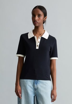 Kobieta ubrana w czarną koszulkę polo z krótkim rękawem, białym kołnierzykiem i mankietami, w połączeniu z jasnoniebieskimi dżinsami, stojąca na jednolitym tle.