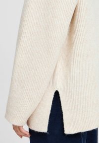 Pull en tricot côtelé crème avec une fente sur le côté et des manches larges, présentant une texture douce et une coupe décontractée, associé à un pantalon bleu foncé.
