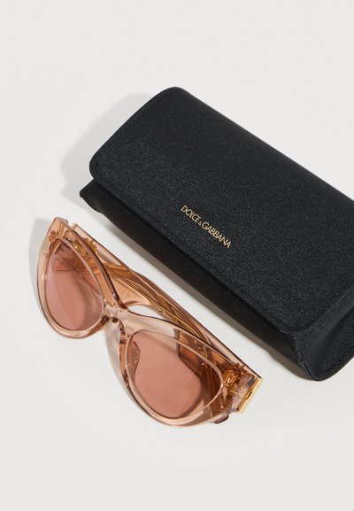 Διαφανή ροζ γυαλιά ηλίου σε σχήμα γάτας δίπλα σε μαύρη υφασμάτινη θήκη Dolce & Gabbana, πάνω σε λευκή επιφάνεια.