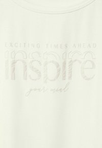 Top in tessuto bianco con testo in rilievo: "TEMPI ECCITANTI IN ARRIVO ispira la tua mente" con un motivo testurizzato sulla parola "ispira".