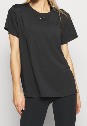 Mujer con una camiseta negra holgada de Nike con el logo blanco en el cuello y leggings negros ajustados, de pie contra un fondo liso.