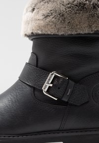 Black leather boot with a faux fur cuff, metal buckle, textured surface, and a padded ankle for comfort. Detailed stitching and logo accent.