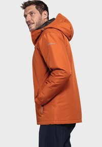 Veste isolante orange avec capuche, équipée d'une poche zippée, de cordons de serrage ajustables et d'une texture lisse. Logo sur la manche gauche.