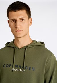 Sudadera verde oliva con un bolsillo tipo canguro en el frente, capucha con cordones y texto en color navy que dice "COPENHAGEN LINDBERGH."