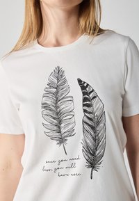 Vit t-shirt i bomull med två handritade svarta fjädersdesigns, tillsammans med ett inspirerande citat i kursiv stil under fjädrarna.