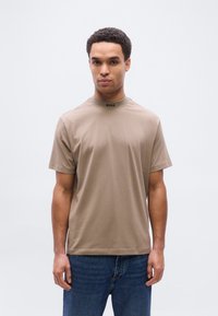 Χακί κοντομάνικη μπλούζα με ψηλό γιακά, με τη λέξη "BOSS" σε μαύρο χρώμα στη λαιμόκοψη· συνδυασμένη με μπλε τζιν.