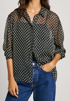 Kobieta nosząca przezroczystą czarną bluzkę zapinaną na guziki z małymi kwiatowymi wzorami, w połączeniu z niebieskimi dżinsami, jedna ręka w kieszeni, druga rozluźniona.