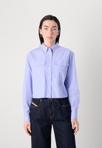 Camicetta corta di colore viola chiaro con colletto a bottoni, due tasche sul petto e maniche lunghe, abbinata a jeans di denim scuro.