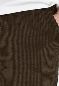 Tessuto di velluto a coste marrone con texture a coste verticali, dotato di tasche e una fascia elastica leggermente elasticizzata. Dettagli di cucitura visibili.