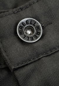 Black fabric close-up featuring a metal snap button with "REELL" embossed, set against a textured background.