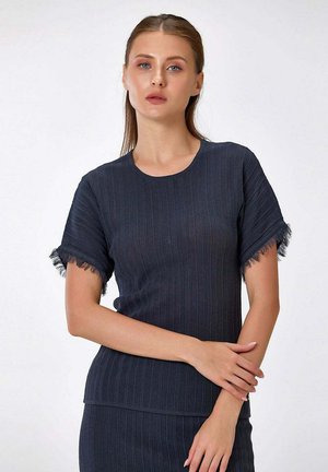Jeune femme aux cheveux châtain clair porte un haut côtelé bleu marine à manches courtes avec des bords de manches à franges, posant devant un fond blanc.