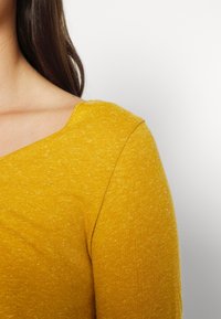 Senapsgul långärmad topp med en strukturerad, något melerad tyg. Har en bred, rundad halsringning och sydda sömmar.