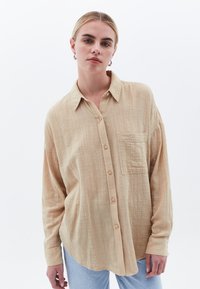 Camicia beige con bottoni realizzata in tessuto testurizzato, dotata di taschino sul petto e maniche lunghe, completata da un orlo arrotondato.