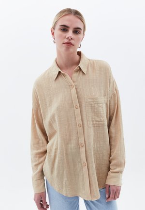 Beige košile na knoflíky vyrobená z texturované tkaniny, s náprsní kapsou a dlouhými rukávy, doplněná zaobleným spodním okrajem.