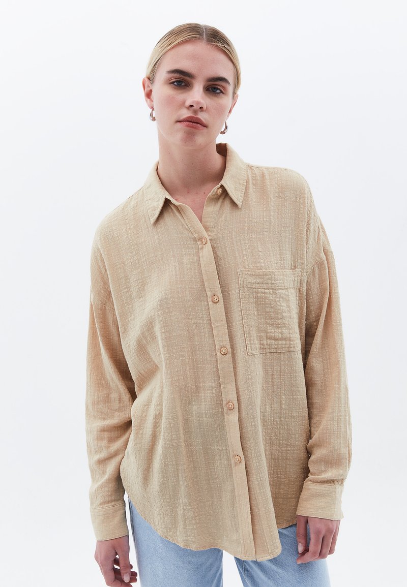 Camisa beige de botón hecha de una tela texturizada, con un bolsillo en el pecho y mangas largas, complementada con un dobladillo redondeado.