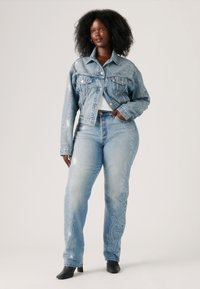 Veste en denim avec broderies blanches décoratives, associée à un jean bleu délavé comportant des broderies similaires. Des bottines noires viennent compléter le look.