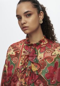 Bunte gemusterte Bluse mit Rüschen, lebhaften Rottönen, Grüntönen und Gelbtönen, mit hohem Ausschnitt und Knopfakzenten. Goldene Ohrringe.