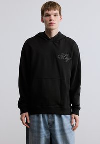 Μαύρο φούτερ με κουκούλα και μπροστινή τσέπη-θήκη, με κεντημένη λεπτομέρεια "Hugo". Κατασκευασμένο από απαλό ύφασμα, σε συνδυασμό με ανοιχτόχρωμο τζιν σορτς.