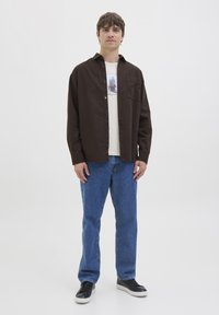 Joven de pie vistiendo una camisa marrón oscuro abotonada sobre una camiseta blanca estampada, jeans azules y tenis negros con suelas blancas.