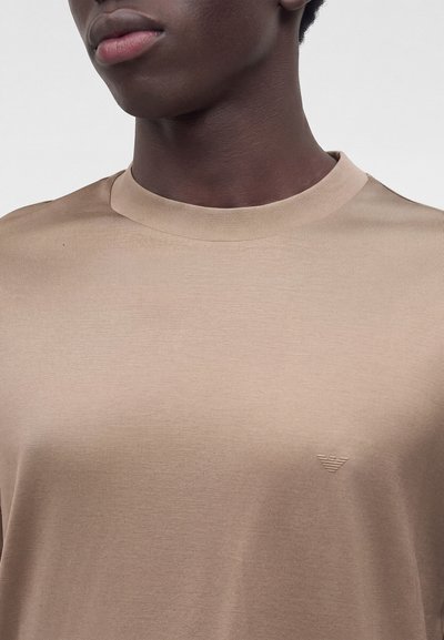 Νεαρό άτομο φοράει μπεζ t-shirt με στρογγυλή λαιμόκοψη και ένα μικρό, διακριτικό λογότυπο στο αριστερό μέρος του στήθους σε απλό φόντο.