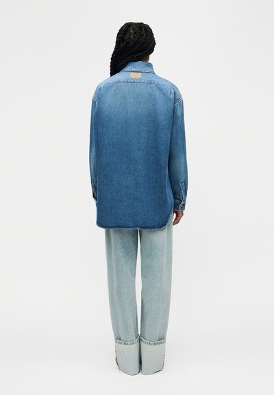Personne aux cheveux longs tressés portant une chemise en jean oversize et un jean bleu clair à jambes larges avec de grands revers, debout de dos.