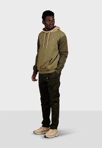 Un sweat à capuche vert olive avec des accents beige, une poche kangourou et des cordons de serrage, associé à un pantalon noir et des baskets de couleur claire.