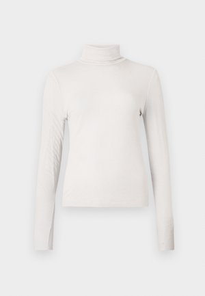 Kremo spalvos raukšlėtas ilgomis rankovėmis golfo tipo marškinėliai su prigludusia siluetu prieš baltą fono.