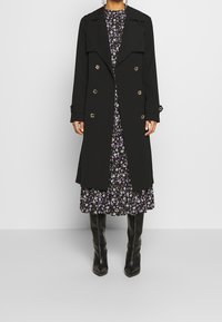 Manteau noir croisé à boutons dorés, col surdimensionné et manches longues, superposé sur une robe imprimée florale et des bottes montant jusqu'aux genoux.
