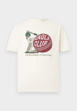 Giocatore di baseball con casco e mazza che colpisce una grande palla rossa con la scritta "Eagle Club" su una t-shirt bianca Emporio Armani.