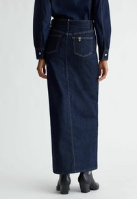 Gonna maxi in denim scuro con design dritto, tasche posteriori e cuciture gialle a contrasto, abbinata a stivaletti neri con tacco.