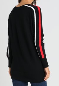 Pull en tricot noir avec une finition texturée, doté de rayures rouges et blanches le long des manches. Manches longues et coupe décontractée.