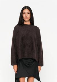 Maglione lavorato a maglia fuzzy marrone scuro con collo alto e maniche lunghe, abbinato a una gonna nera asimmetrica fluida.