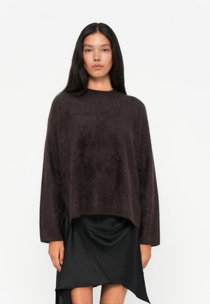 Maglione lavorato a maglia fuzzy marrone scuro con collo alto e maniche lunghe, abbinato a una gonna nera asimmetrica fluida.