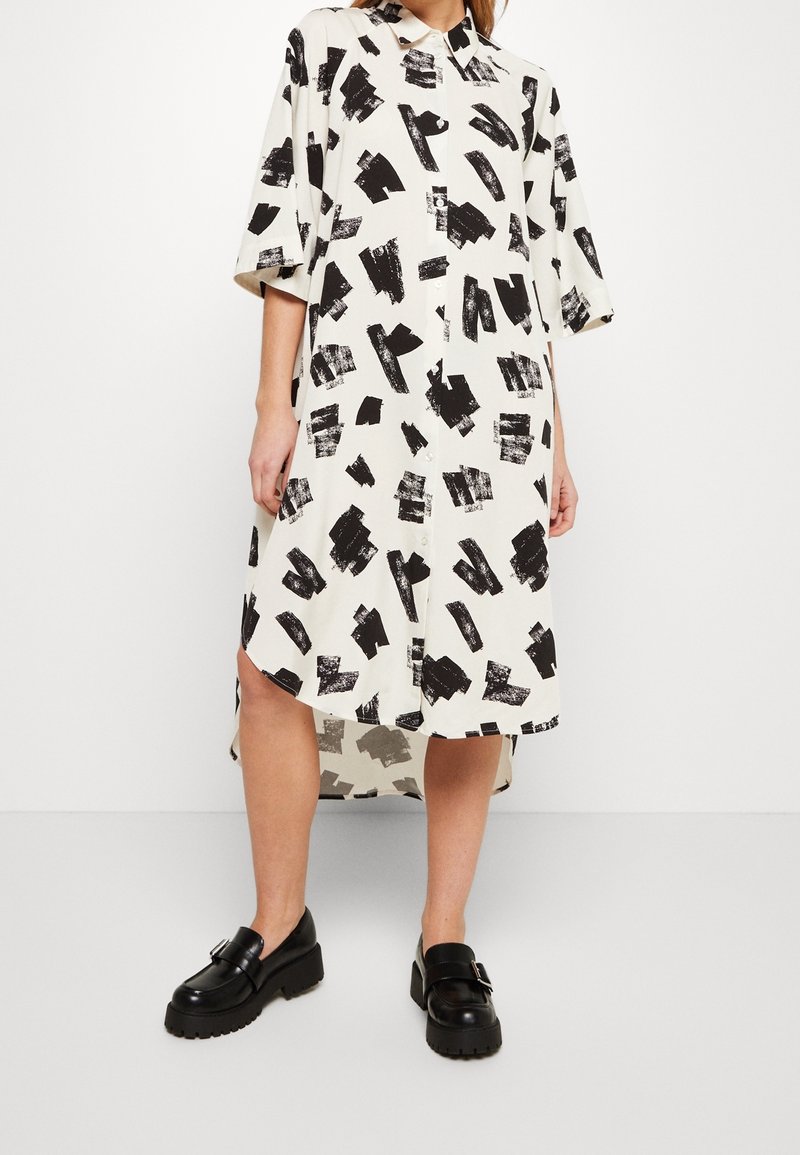 Femme portant une robe chemise midi blanche avec des motifs à coups de pinceau noirs, demi-manches, boutons sur le devant, associée à des mocassins noirs épais.