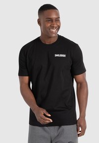 Sort bomulds t-shirt med korte ærmer, der har den hvide tekst "SMILODOX" på venstre brystområde. Modellens påklædning inkluderer grå shorts.