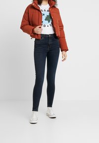 Chaqueta acolchada roja con cuello alto, combinada con una camiseta gráfica blanca y jeans ajustados de color azul oscuro. Zapatillas blancas completan el conjunto.