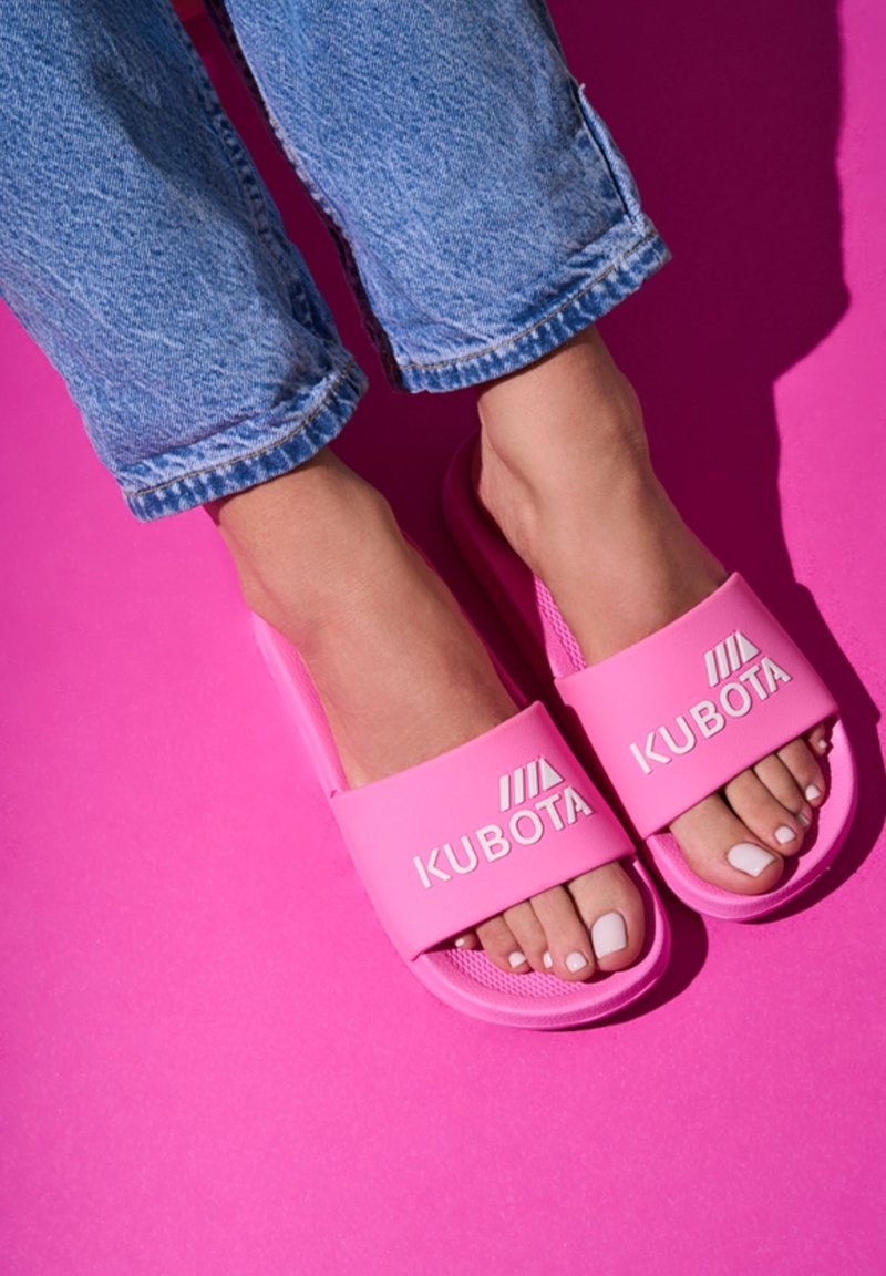 Pink slip-on sandals with a textured sole and a wide strap featuring the "KUBOTA" logo. Paired with light blue jeans against a pink background.