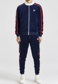 Marinblå velourträningsoverall med vita och burgundyröda detaljer, bestående av en zip-up jacka och byxor med elastisk midja, logga på vänster bröst och lår.