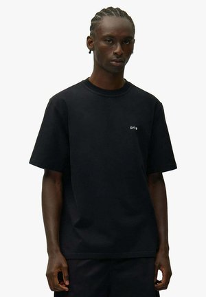 Giovane con capelli intrecciati che indossa una maglietta nera a maniche corte con un piccolo logo bianco "arte" sul petto a sinistra, in piedi contro uno sfondo bianco.