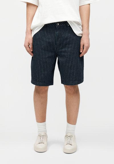 Άνδρας που φοράει ναυτικά μπλε σορτς με ρίγες μέχρι το γόνατο, λευκές κάλτσες, λευκά αθλητικά παπούτσια και μια άνετη λευκή μπλούζα, στέκεται σε λευκό φόντο.