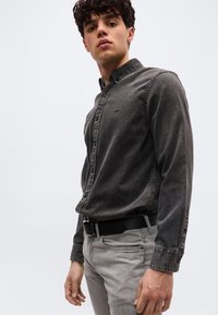 Νέος άνδρας με σγουρά μαλλιά που φοράει ξεθωριασμένο μαύρο πουκάμισο με κουμπιά, μπαλωμένο μέσα σε ανοιχτόχρωμο γκρι παντελόνι με μαύρη ζώνη, πάνω σε απλό φόντο.