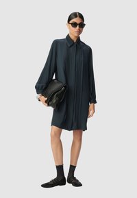Vestito strutturato blu navy con dettagli plissettati e scollo con colletto, abbinato a una borsa a tracolla in pelle nera e scarpe flat con calzini alla caviglia.