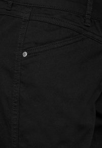 Schwarze Denim-Chinos mit glatter Textur, ausgestattet mit einer vorderen Tasche mit silbernem Knopf und tonaler Naht für ein nahtloses Design.