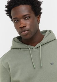 Młody mężczyzna z krótkimi kręconymi włosami, ubrany w jasnoróżową bluzę z kapturem, z sznurkami i małym logo konia na piersi.