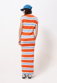 Gestreiftes, ärmelloses Maxikleid in Orange, Blau und Creme. Weiches Strickmaterial, figurbetonte Silhouette, auffällig durch die horizontalen Streifen. Silberne Schuhe.