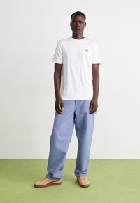 Muž stojící na zelené podlaze, oblečený v bílém tričku, světle modrých volných kalhotách a béžových teniskách s oranžovým pruhem, na pozadí bílé zdi.