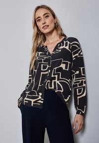 Blusa nera e beige con motivo geometrico astratto, maniche lunghe, scollatura a V e chiusura con bottoni, abbinata a pantaloni blu navy.