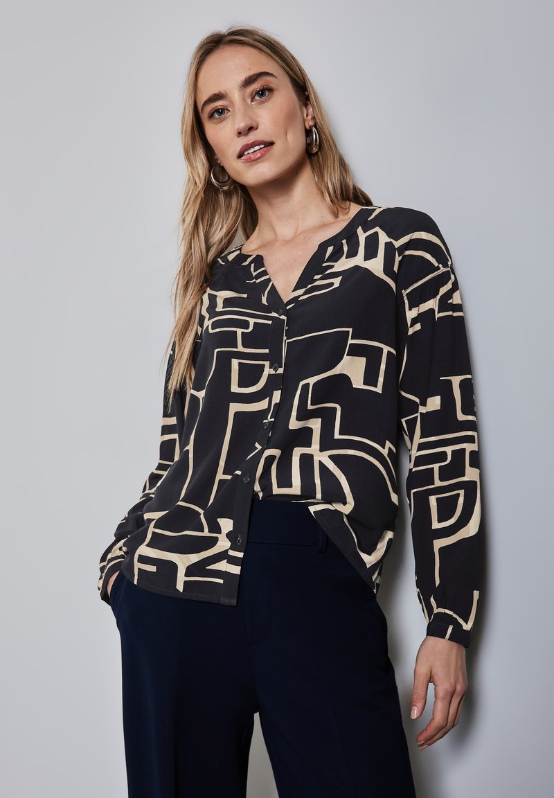 Blusa nera e beige con motivo geometrico astratto, maniche lunghe, scollatura a V e chiusura con bottoni, abbinata a pantaloni blu navy.