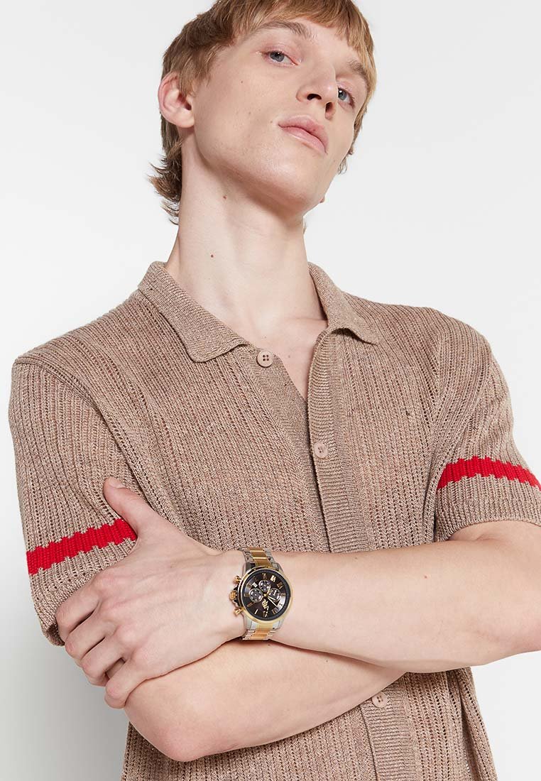 Polo de punto beige con mangas cortas, que presenta una franja roja en la manga izquierda. Se lleva un reloj de pulsera negro y dorado en la muñeca.