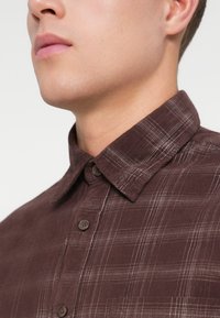 Brun rutete skjorte med strukturert krage, knappestolpe foran og en teksturert stoff. Har subtile vertikale striper og brune knapper.